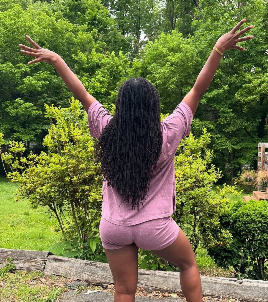 Back view of a woman with long black locs standing outdoors in a garden with her arms raised