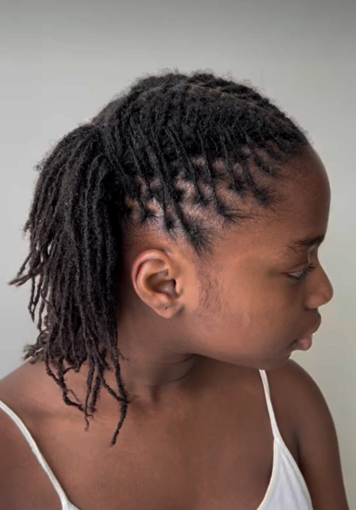 Side view of a woman with neat starter locs, showing clean parts, budding texture, and locs pulled into a low ponytail.